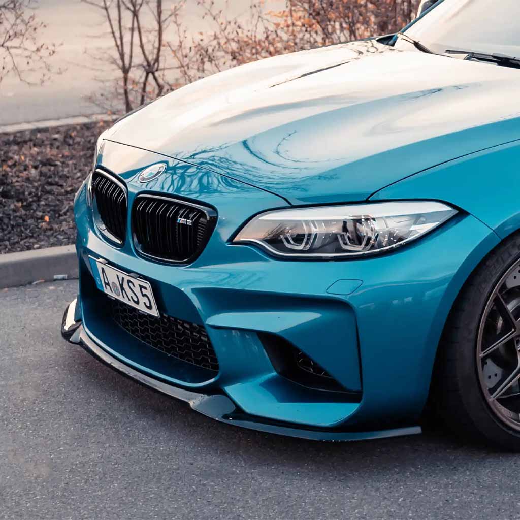 Nahaufnahme der Front eines blauen BMW F87 N55 mit einer ZAERO Design EVO-S Frontlippe von ZAERO, geparkt auf einer Straße in der Nähe von Büschen.