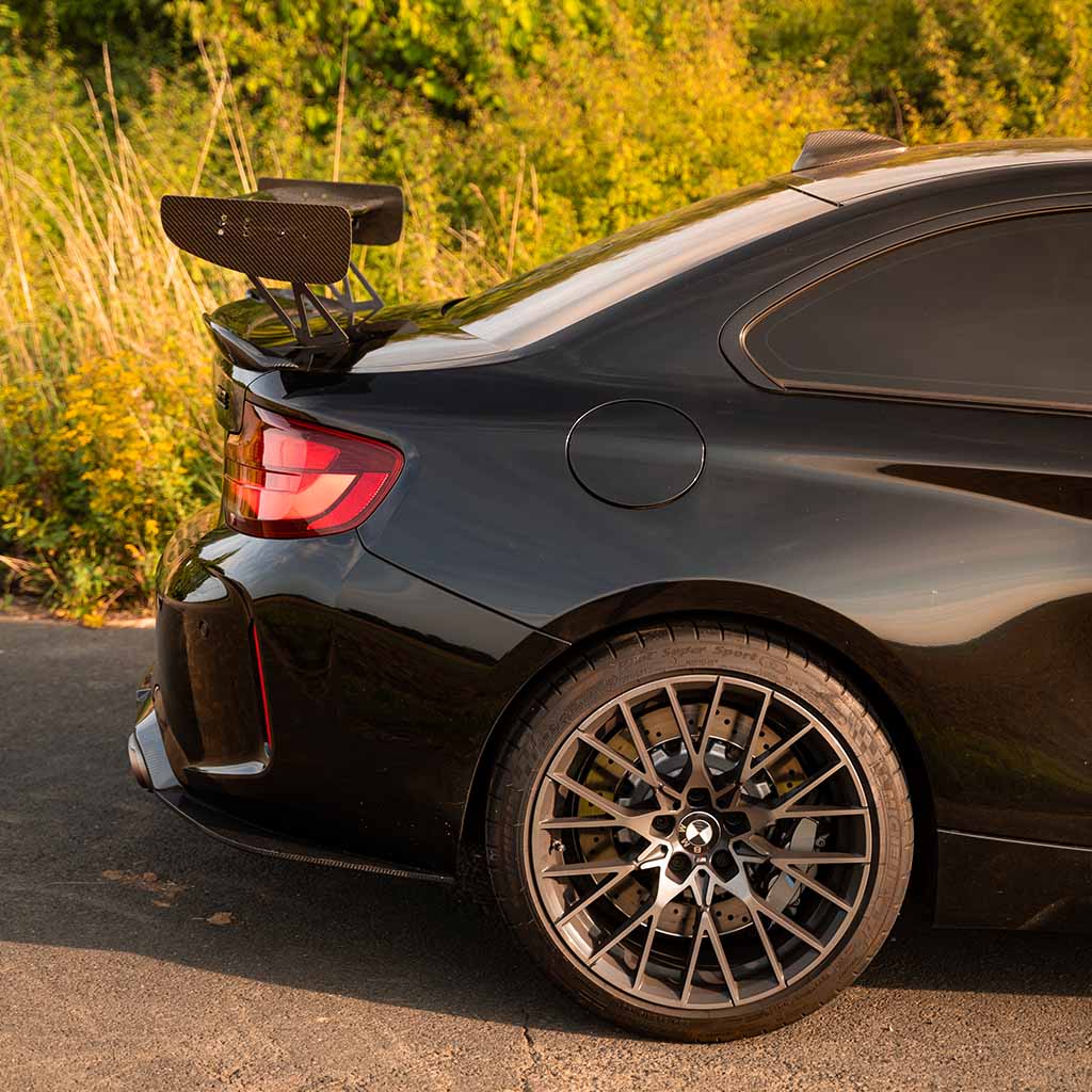 Schwarzer BMW M2 Competition mit einem 55Parts Motorsport Carbon Heckspoiler Heckflügel inklusive Teilegutachten, getönten Scheiben und Leichtmetallfelgen im Grünen geparkt.