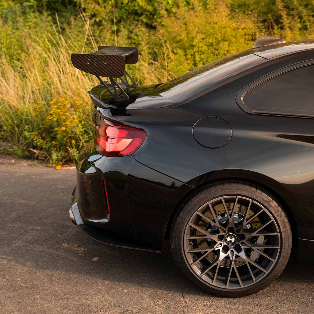 Ein schwarzer BMW M2 Competition durch hohes Gras, der am Heck einen 55Parts Motorsport Carbon Heckspoiler Heckflügel (INKL. Teilegutachten), sowie das Hinterrad, Rücklicht und einen Teil der Heckscheibe zeigt.