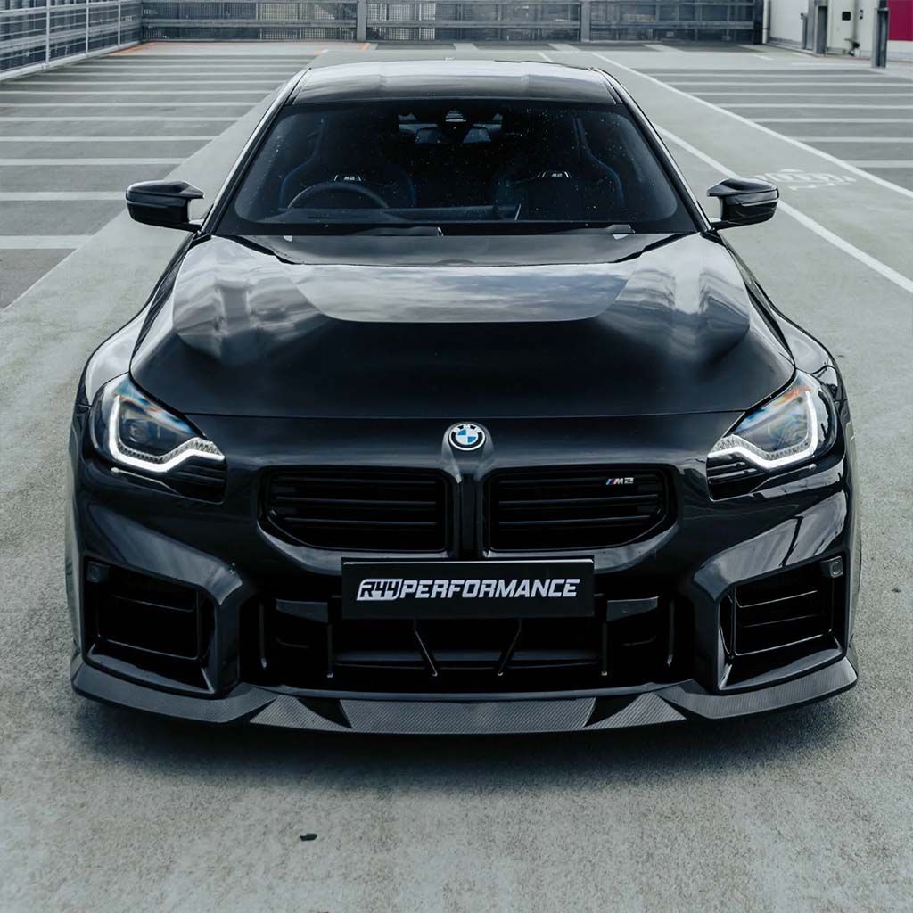 Frontansicht eines schwarzen BMW M2 (G87) mit dem Twistnfix MHC TNF+ Edition 1 Frontspoiler Carbon, inklusive Teilegutachten, geparkt auf einem leeren überdachten Parkplatz.