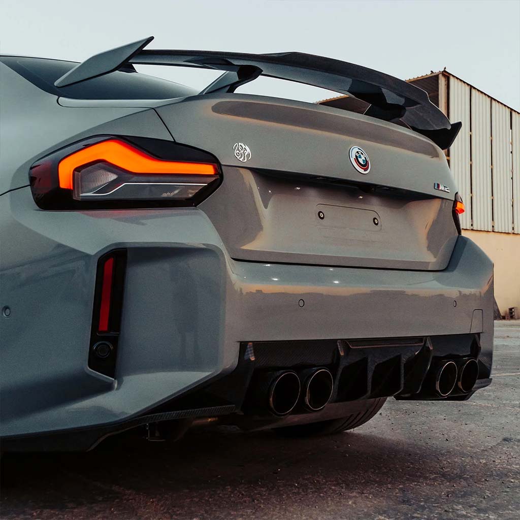 Grauer BMW M2 (G87) mit Twistnfix MHC TNF+ Edition 1 Heckdiffusor Carbon, großem Spoiler, vierfachem Auspuff und scharfen Rückleuchten, geparkt in der Nähe eines Industriegebäudes.
