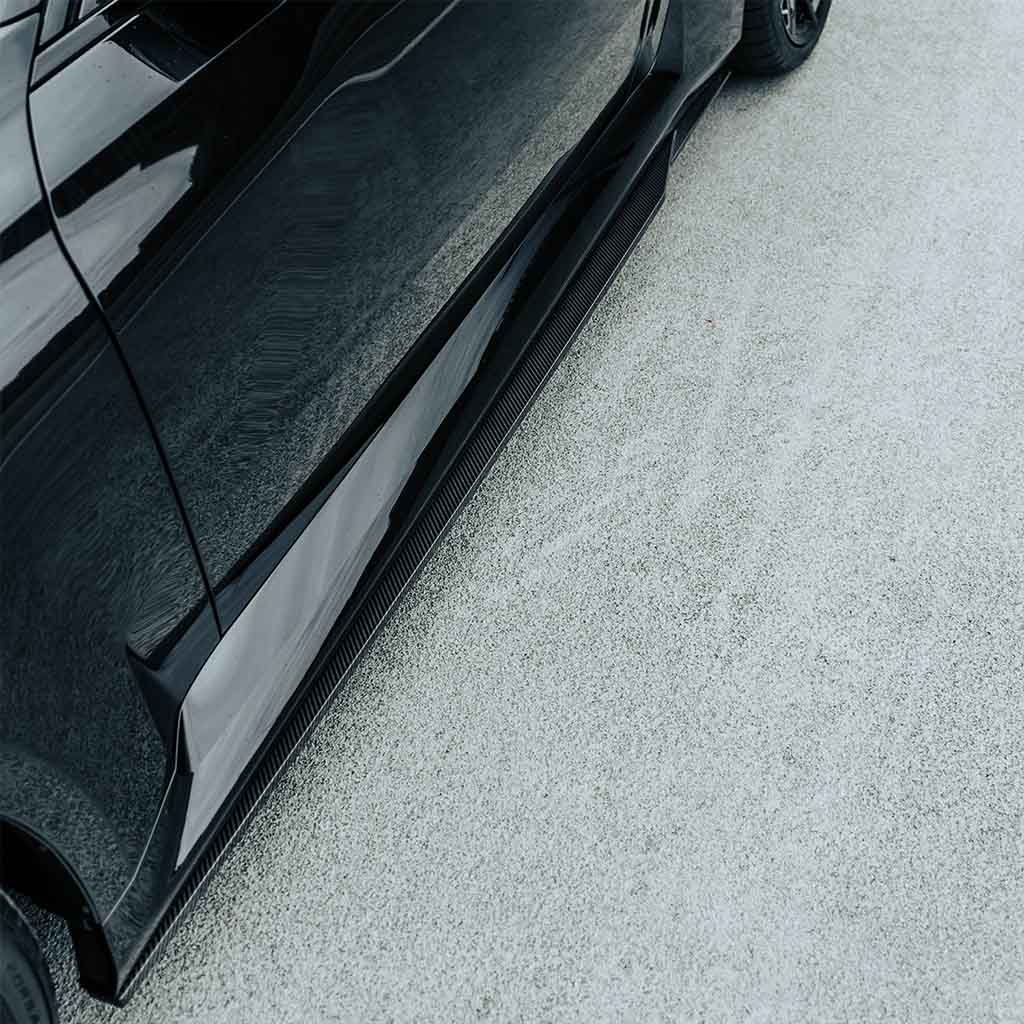 Nahaufnahme von Twistnfix MHC TNF+ Edition 1 Seitenschweller Carbon für BMW M2 (G87) inkl. Teilegutachten, Hervorhebung der Seitenschweller und Reifen auf einer hellgrauen strukturierten Oberfläche.