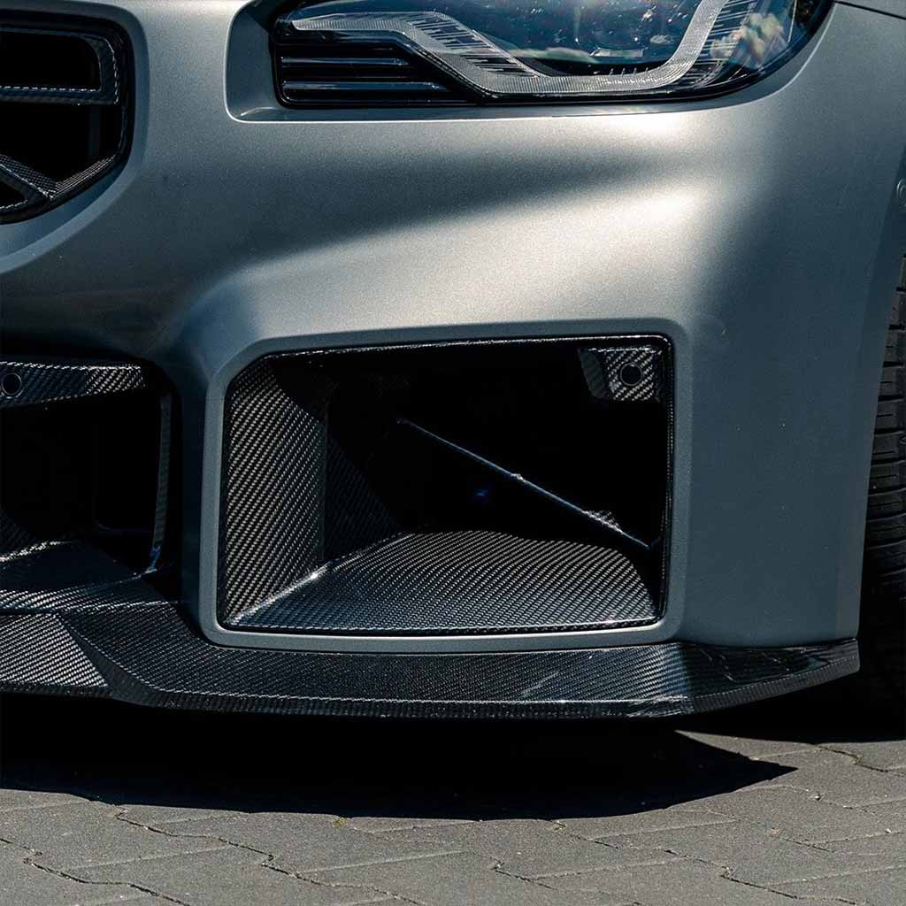 Nahaufnahme eines BMW M2 (G87) Frontstoßfängers mit Twistnfix TNF+ Lufteinlässe Carbon inklusive Teilegutachten und einem Prepreg-Kohlefaser-Splitter, geparkt auf dem Bürgersteig.