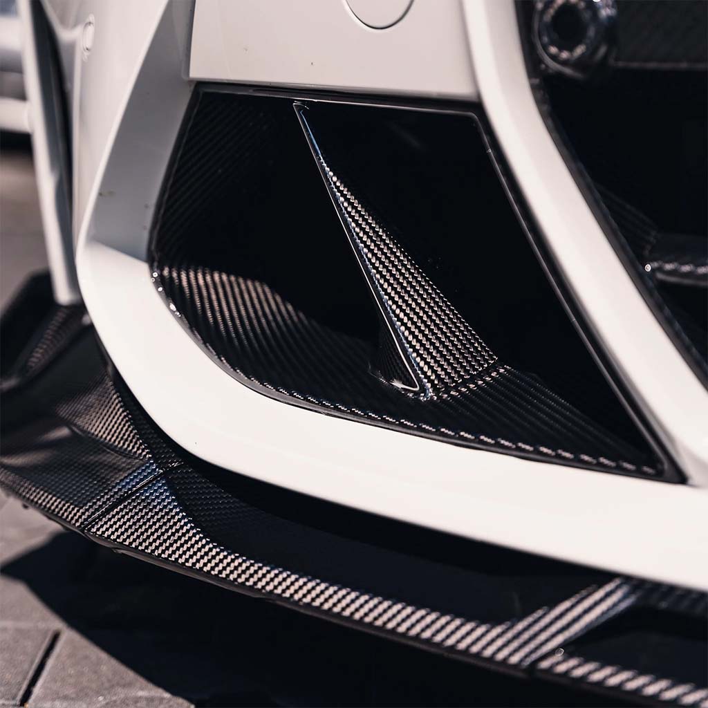 Nahaufnahme von Twistnfix TNF+ Performance Lufteinlässe Carbon für BMW M3 (G80/G81) &amp; M4 (G82/G83), mit eleganten Kohlefaserdetails und Teilegutachten, abgebildet auf einem weiß lackierten Stoßfänger.