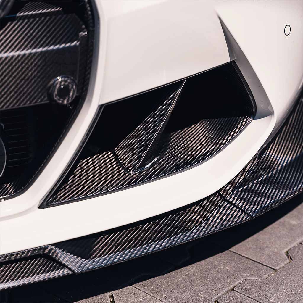 Nahaufnahme der Frontstoßstange eines weißen Fahrzeugs mit Twistnfix TNF+ Performance Lufteinlässe Carbon für BMW M3 (G80/G81) &amp; M4 (G82/G83), mit Prepreg-Carbon-Details und sportlichem Design, inkl. Teilegutachten.