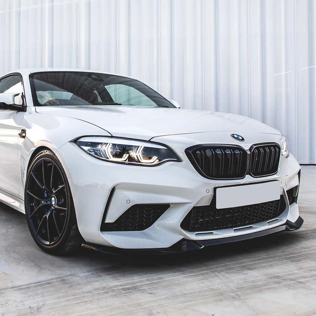 Ein weißer BMW F87 Competition mit der ZAERO Design EVO-S Frontlippe von ZAERO ist auf einer Betonfläche vor einem modernen weißen Hintergrund geparkt.