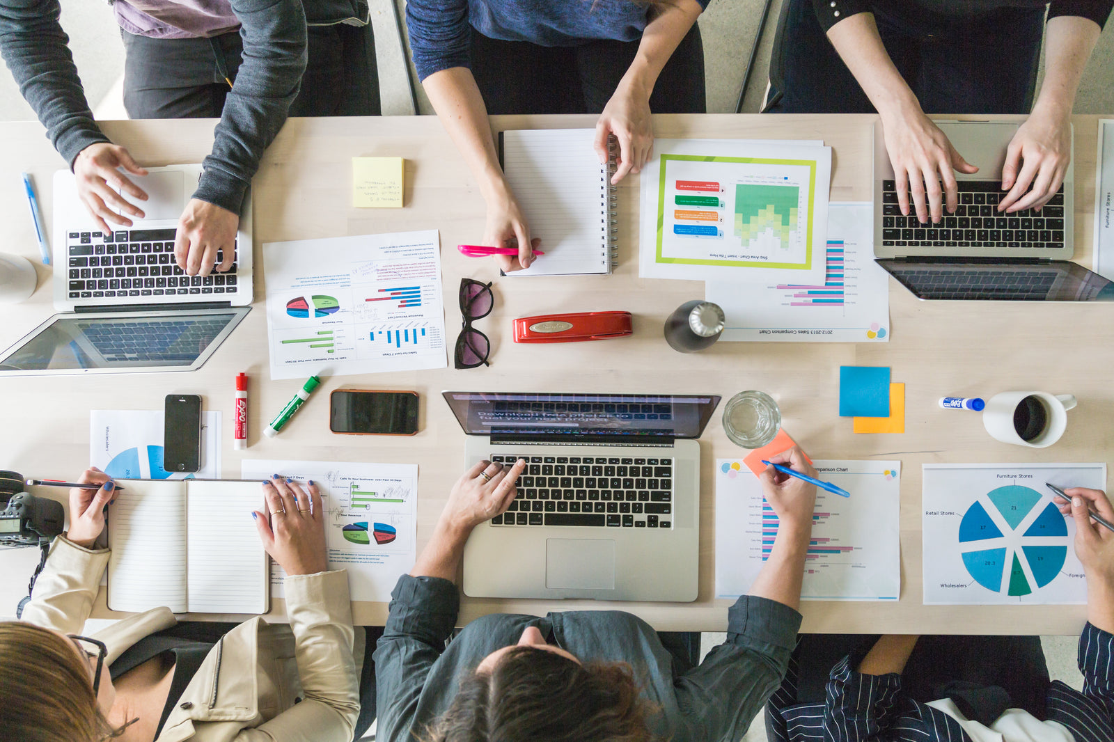 Menschen arbeiten an einem Tisch zusammen, an dem Laptops, Notizbücher, Diagramme und Grafiken verstreut herumliegen, während sie Daten analysieren.