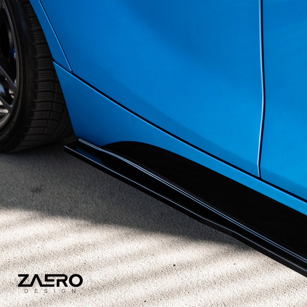 Nahaufnahme der ZAERO Evo-1 Seitenschweller passend für BMW F2x in Glanz Schwarz von ZAERO, dargestellt auf einem blauen Auto, das auf hellem Beton geparkt ist. Das Zaero Design-Logo ist sichtbar.